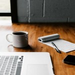 Desk with a notepad, a coffee cup, laptop.